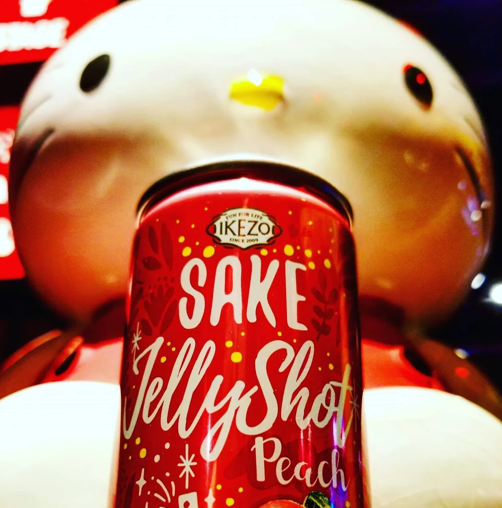 Red SAKE JellyShot Peach can, blurred white feline character in dim light.