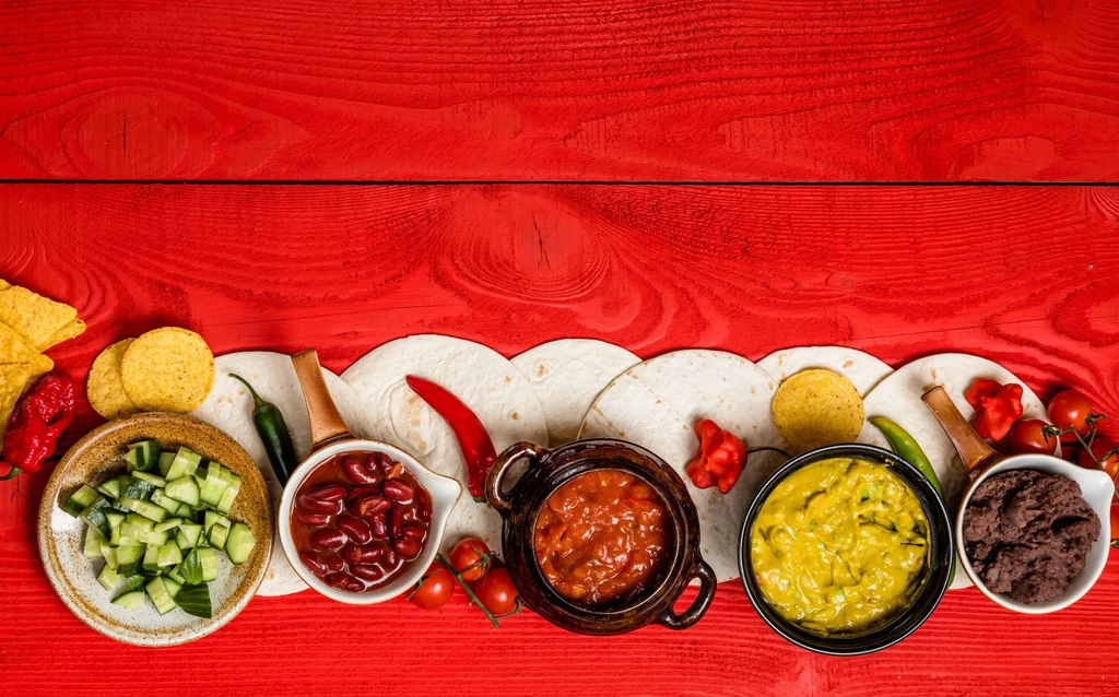 Top-down view of colorful Mexican dishes and ingredients on red wood.