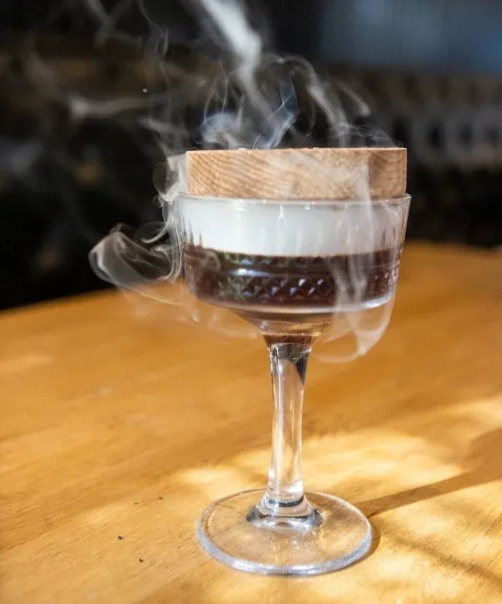 Smoked cocktail: dark liquid, white layer, wooden cap, swirling smoke.