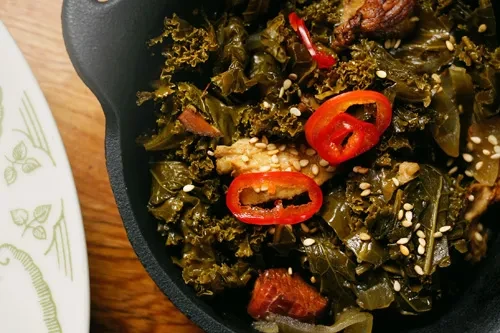 Close-up of stir-fried greens, sesame, chili in dark bowl on wood.