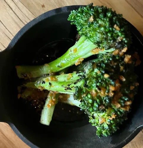 Overhead view of charred broccolini in a dark bowl with crispy toppings.
