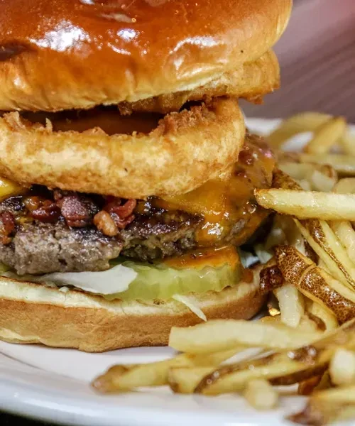Stacked burger with onion ring and skin-on fries on white plate.