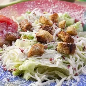 Salad close-up: lettuce, croutons, cheese, tomato, red seasoning on speckled plate.