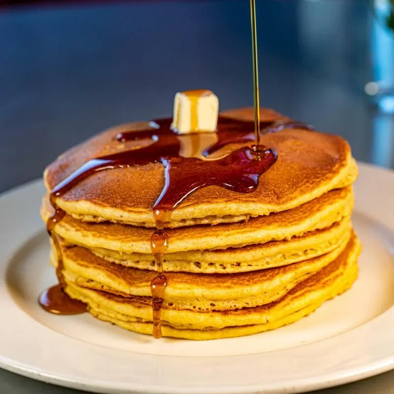 Stack of golden-brown pancakes with melting butter and syrup being poured.