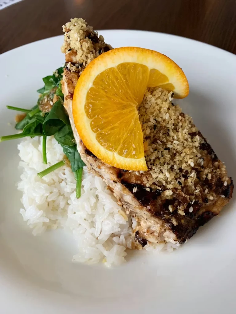 Top-down view of seared fish/chicken with sesame topping, orange, rice, greens.