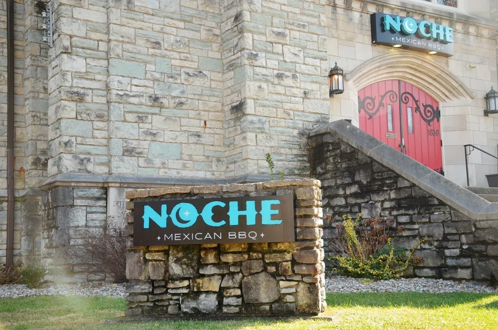 Stone building, NOCHE + MEXICAN BBQ, with prominent turquoise signs and arched entrance.