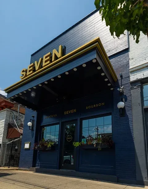 Modern dark 'SEVEN' bar exterior, gold lettering, under clear blue sky.