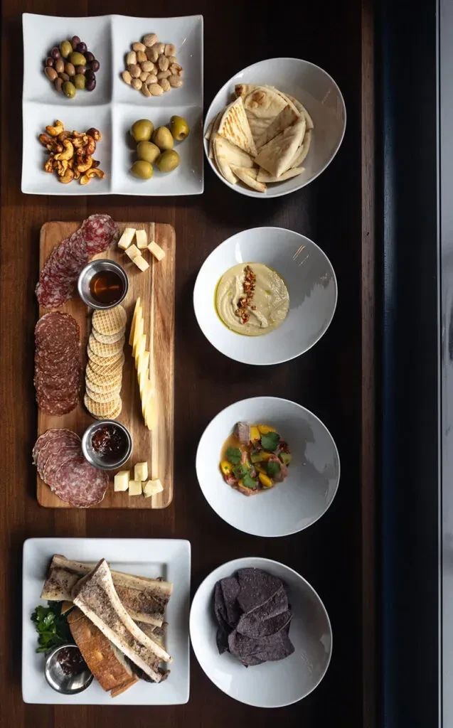 Dark table laden with diverse small plates of charcuterie, ceviche, and dips.