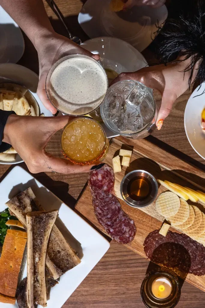 Top-down, four hands clink diverse drinks at a restaurant table.