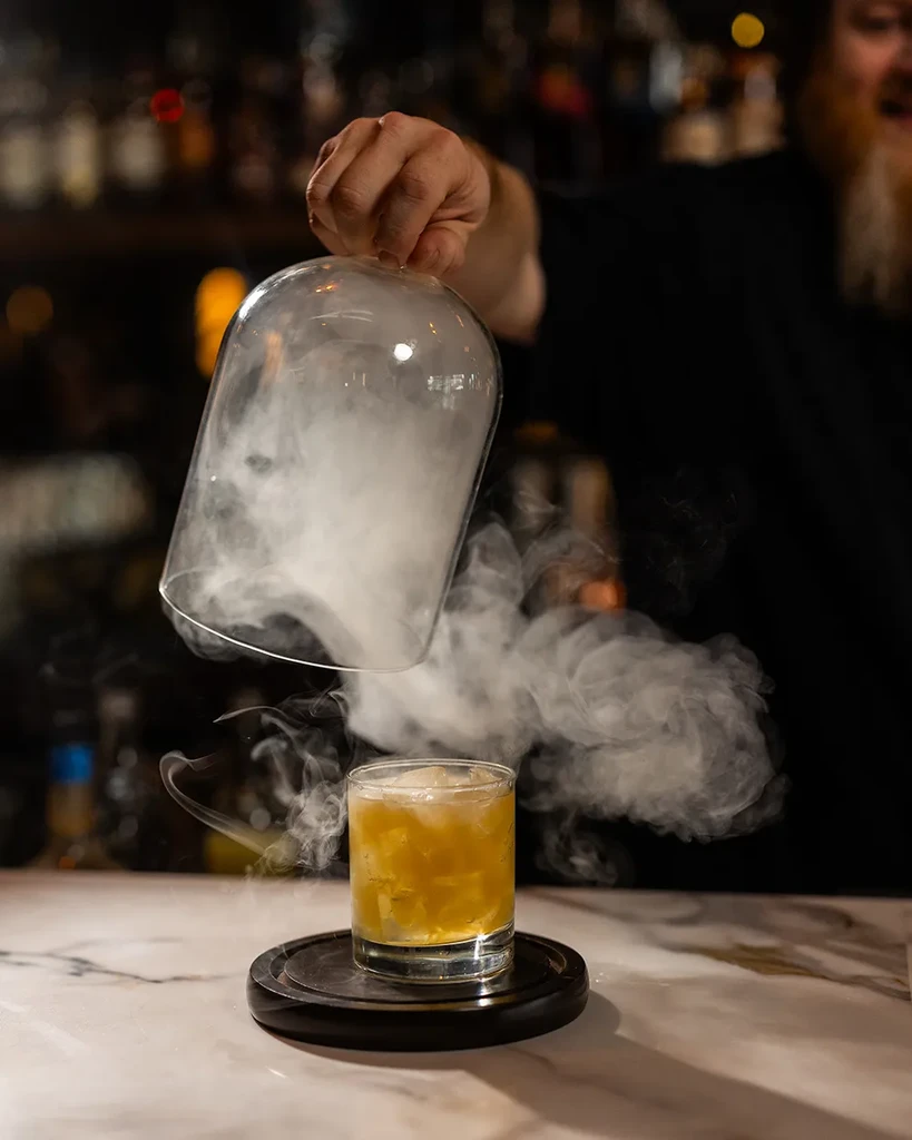 Hand lifts cloche, smoke billows around cocktail on marble bar.