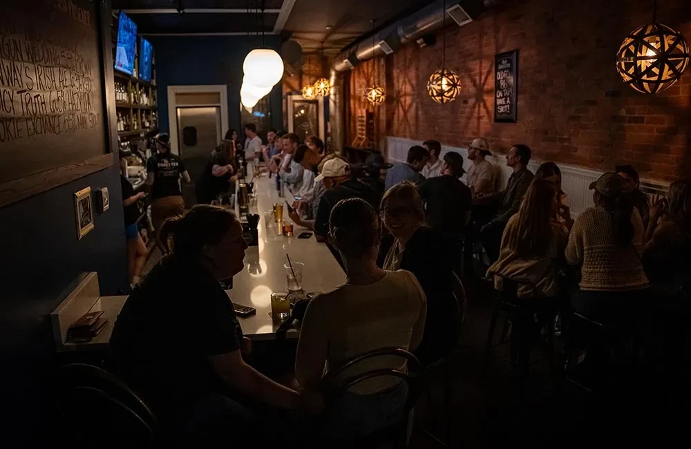 Warm, dimly lit bar interior with patrons, a long bar, and brick wall.