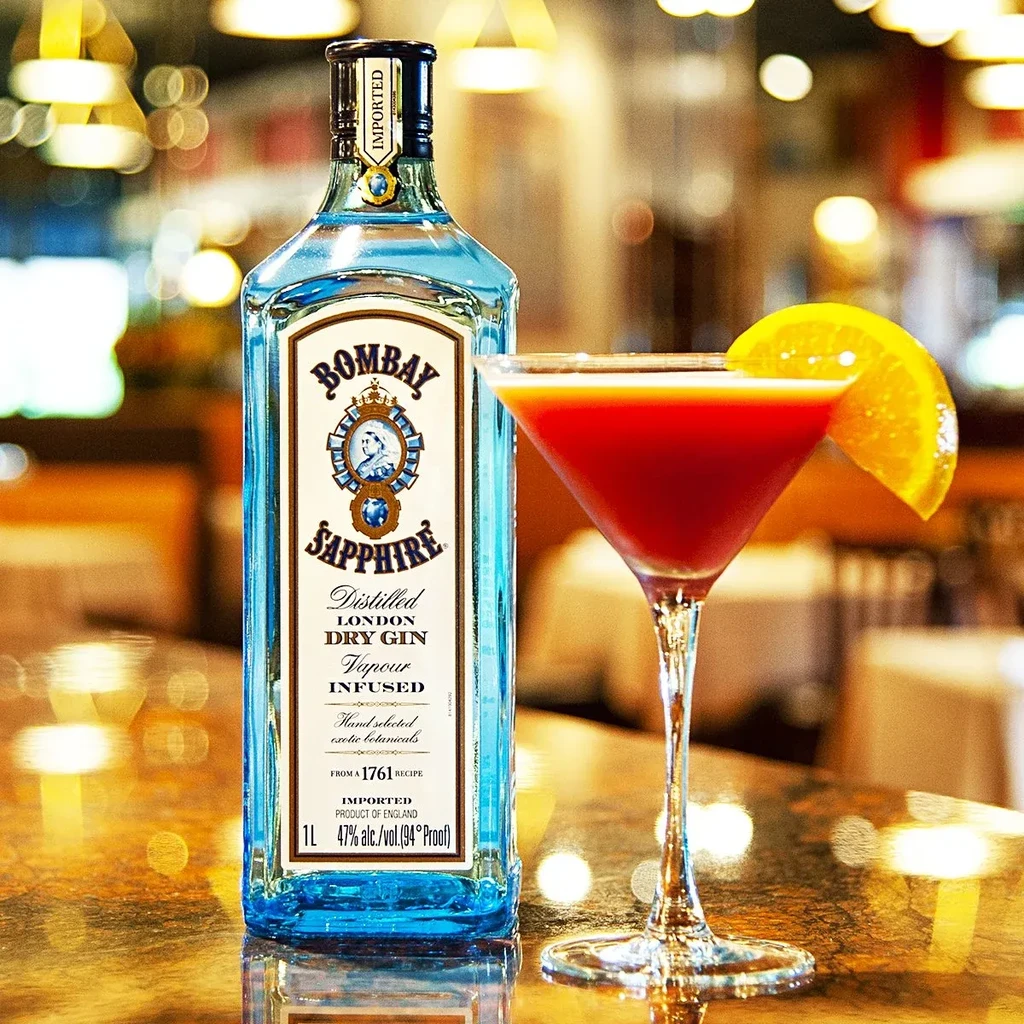 Bombay Sapphire gin bottle next to a red cocktail with orange slice.