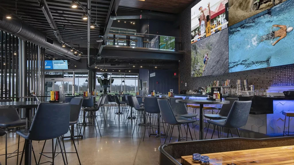 Modern Topgolf venue interior: sports screens, bar, dining, and golf range.
