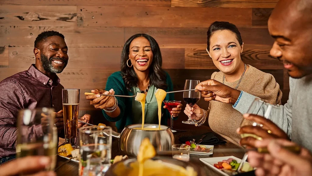 Four friends laughing, sharing fondue with cheese dripping from forks.