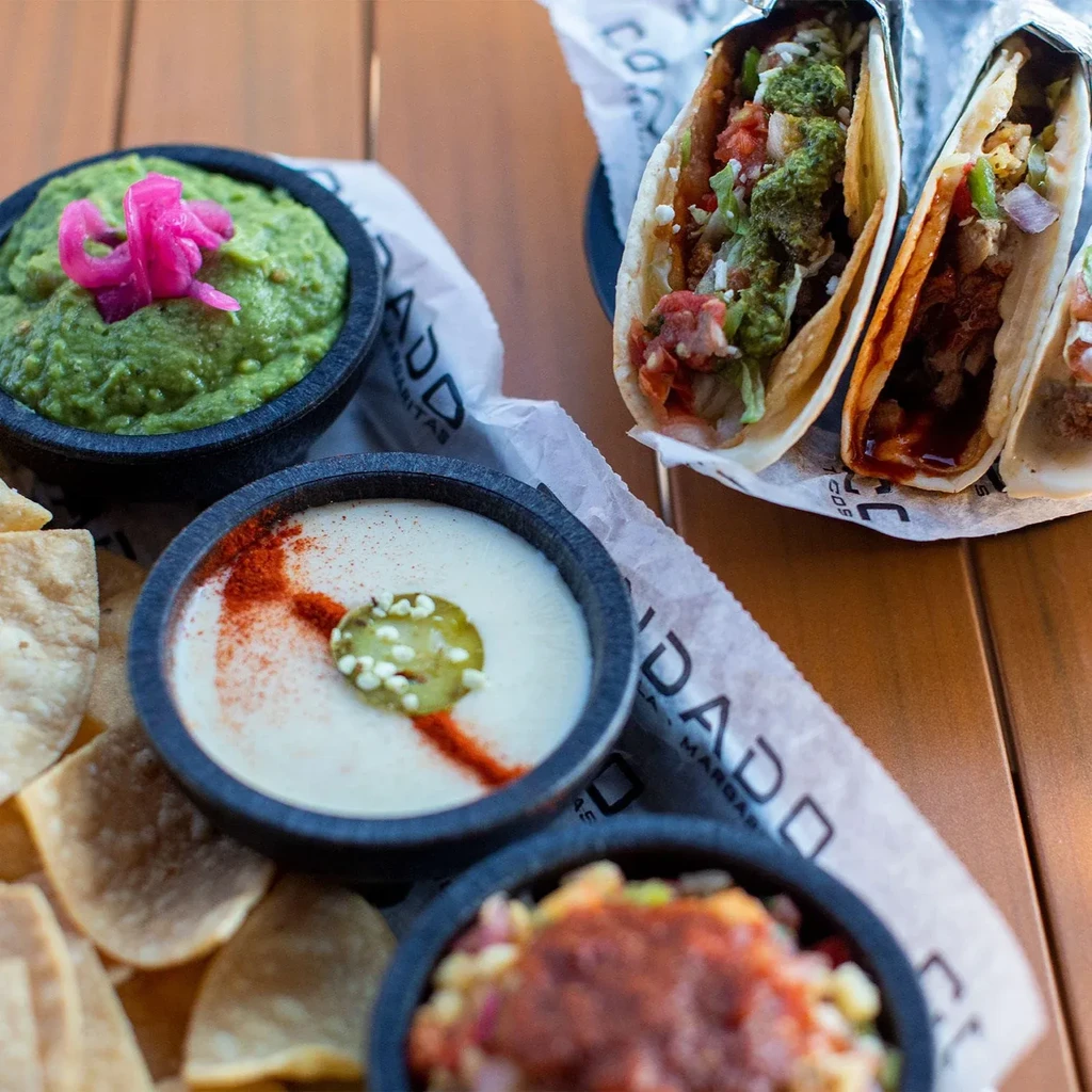 Mexican food dishes: guacamole, queso, tacos, chips on wooden table.