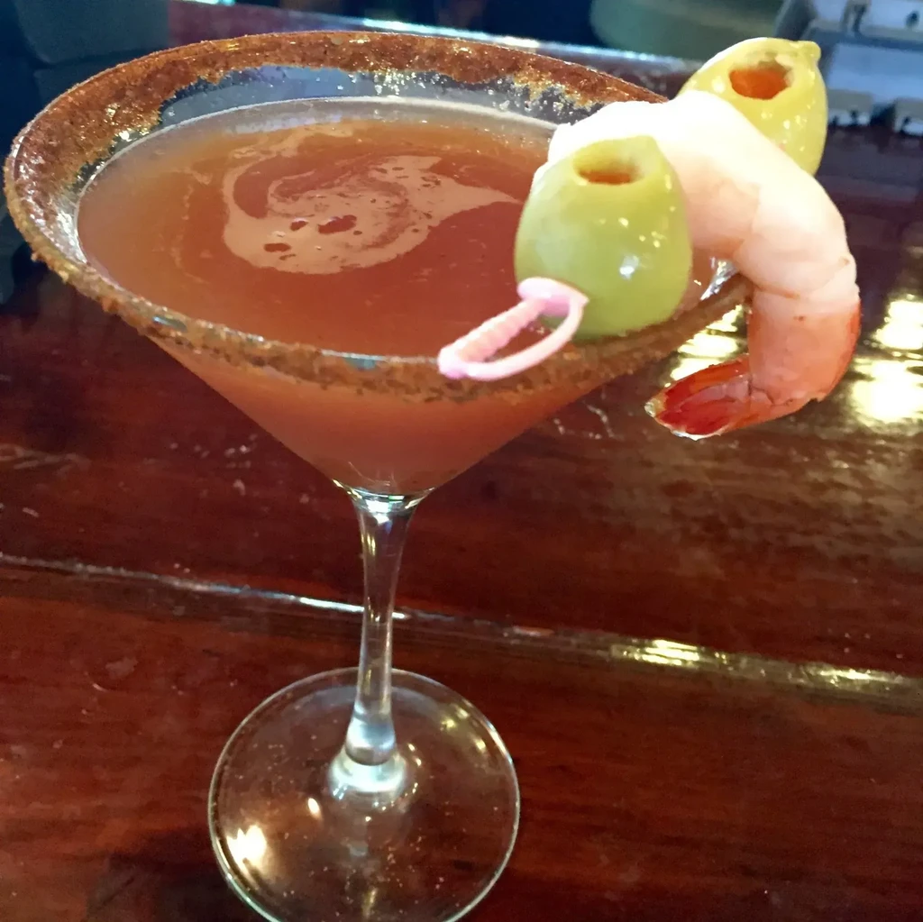 Reddish-brown cocktail garnished with shrimp, olives, and spiced rim.