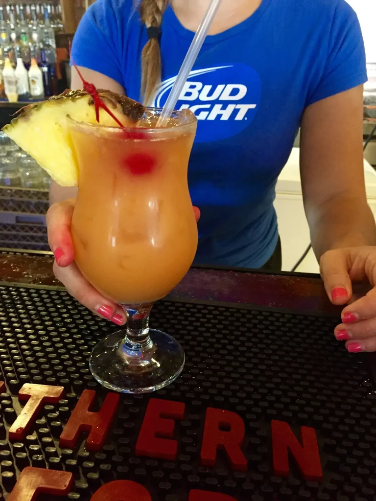 Person with pink nails and Bud Light shirt holds an orange cocktail.