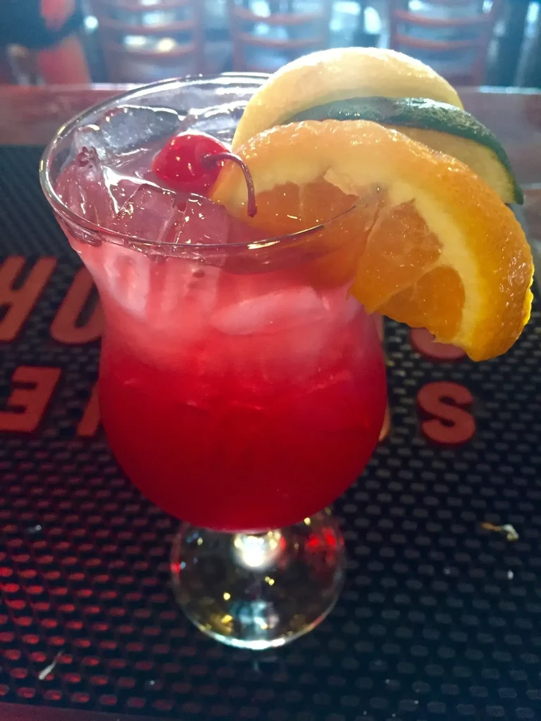 Vibrant red cocktail in hurricane glass, garnished with fruit and ice.