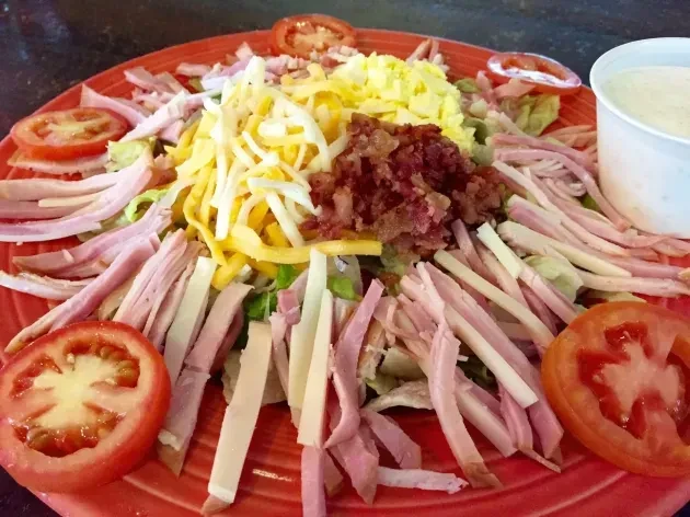 Cobb salad with cheese, bacon, ham, tomato, dressing on red plate.
