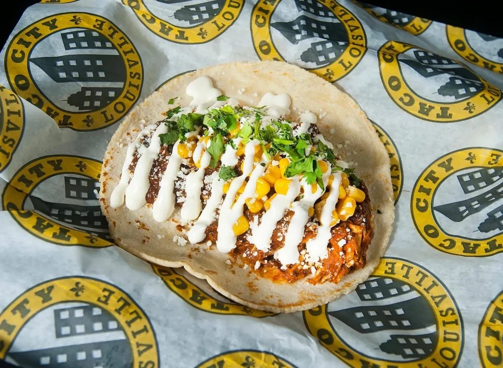 Single taco with dark filling, corn, cotija, crema, and cilantro.
