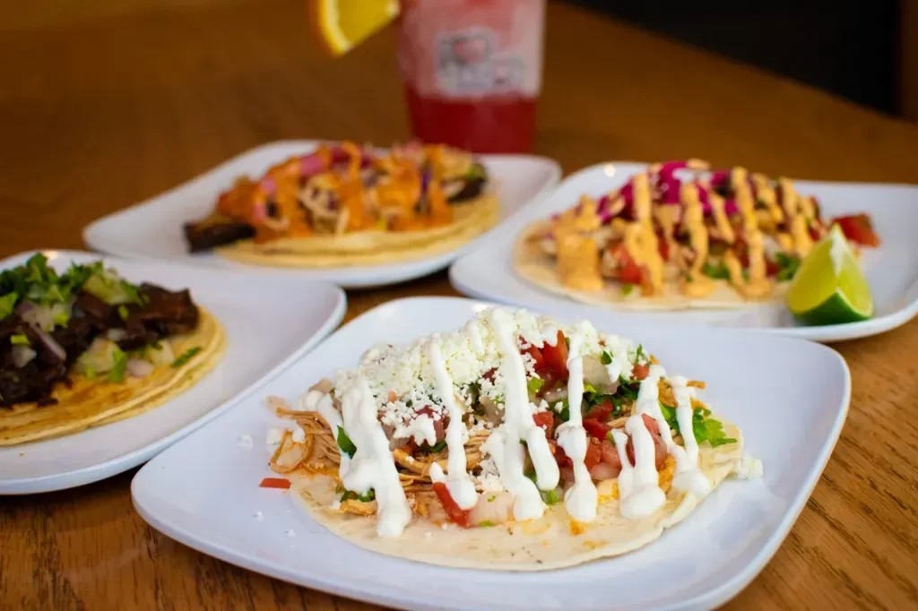 Four plates of various tacos and a drink on a wooden table.