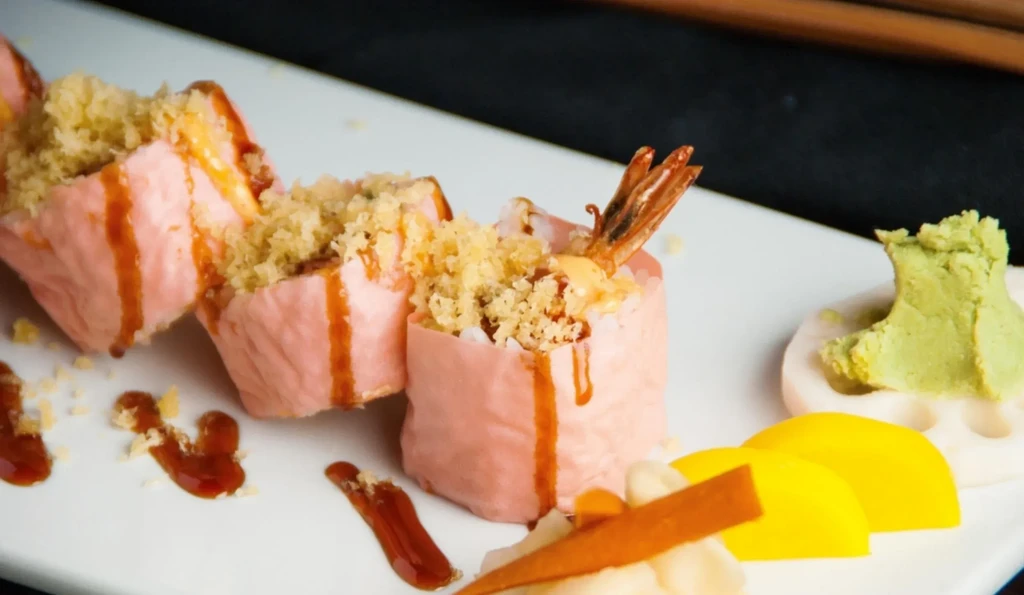 Pink-wrapped shrimp tempura sushi rolls with flakes, sauce, and garnishes.