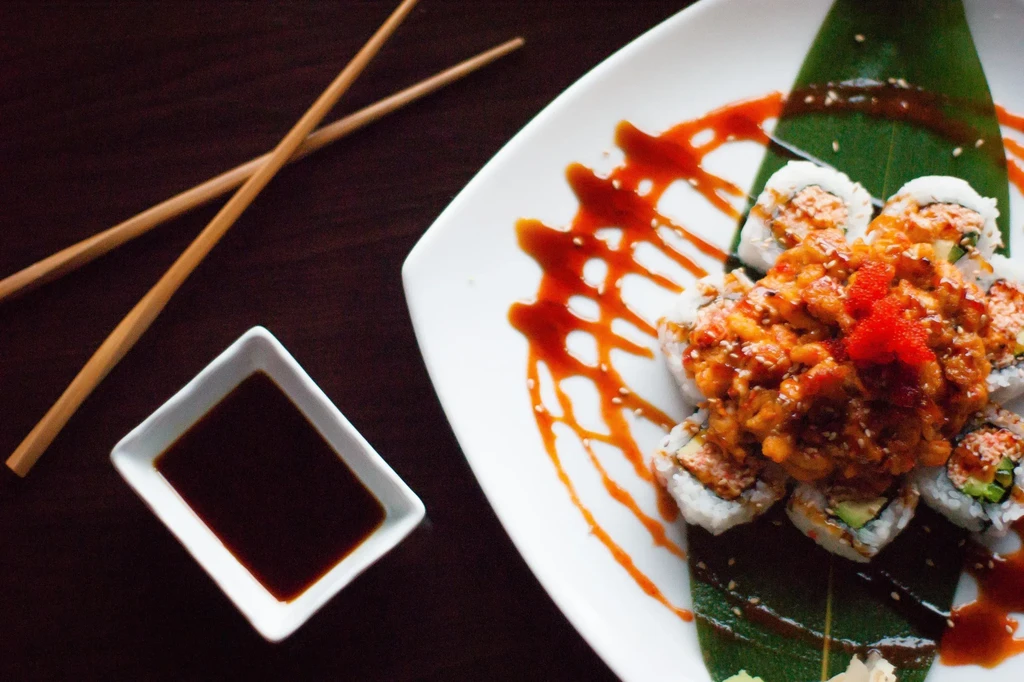 Flower-shaped sushi roll with spicy topping, sauce, and chopsticks.