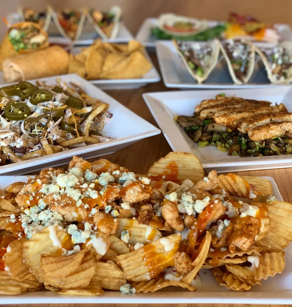 Table of buffalo chicken nachos, loaded fries, grilled chicken, and other dishes.