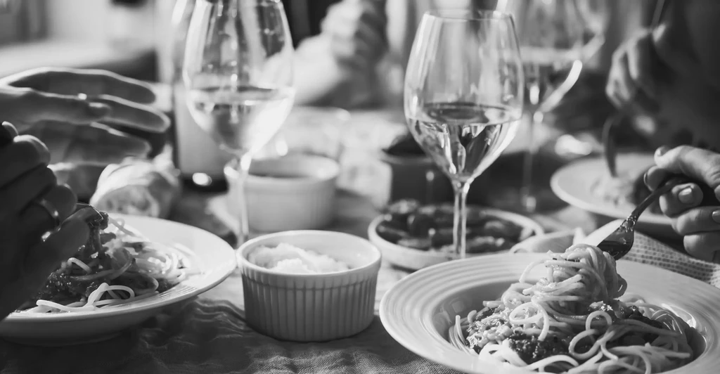 Black and white close-up of hands eating spaghetti and drinking wine.