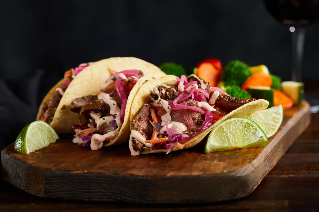 Three steak tacos, cabbage, pickled onions, sauce, limes, on wooden board.