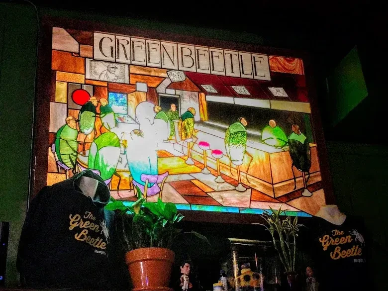 Illuminated stained-glass of beetles at a bar, 'GREENBEETLE', above branded shirts.