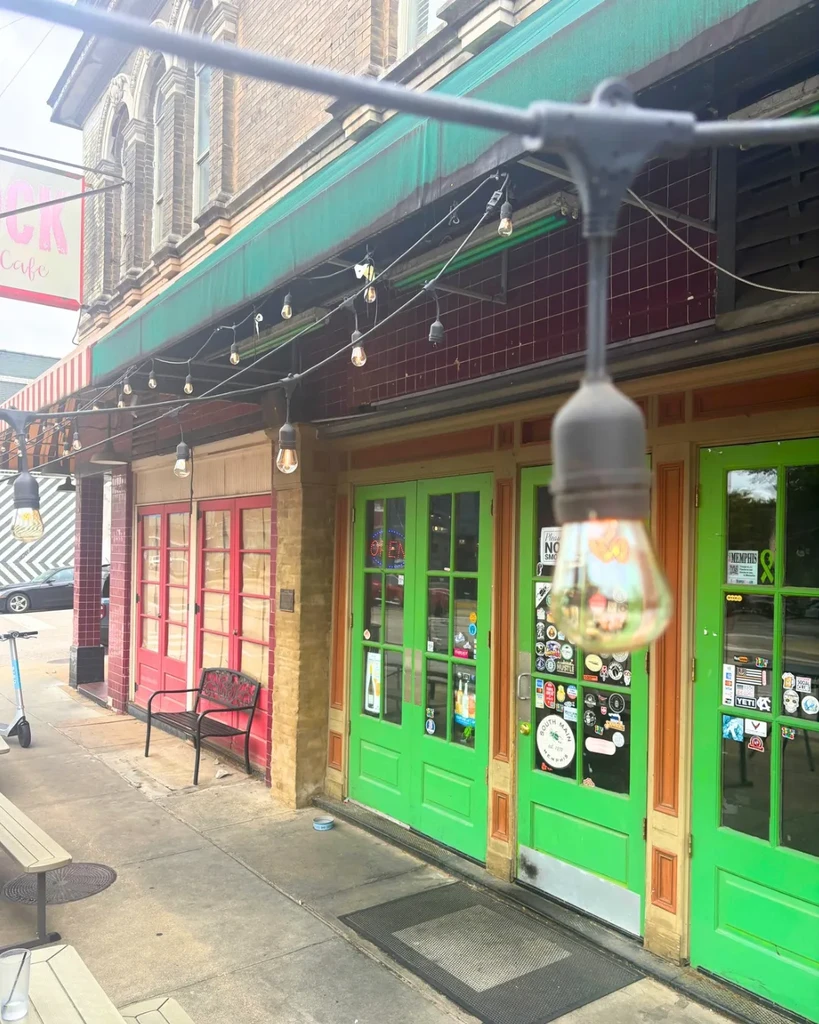 Restaurant exterior with colorful doors, green awning, string lights, and benches.