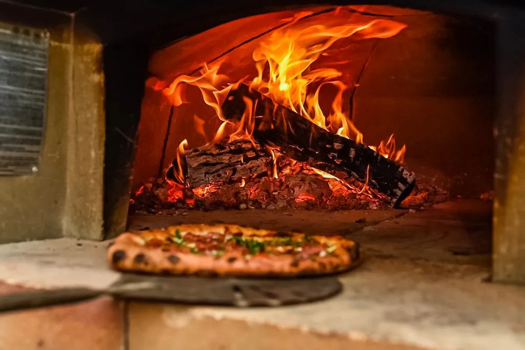 Pizza cooking inside a brick wood-fired oven with roaring flames.
