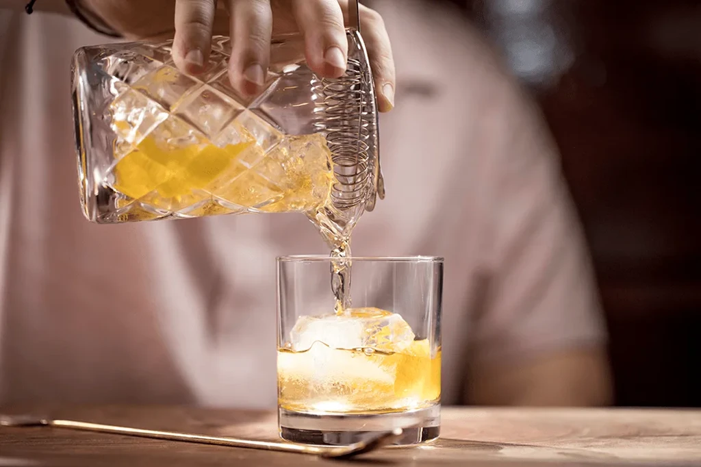 Bartender's hand pours golden-amber liquid from strainer into glass.
