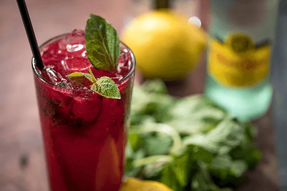 Red drink with ice, mint, and straw; blurred lemon and bottle background.