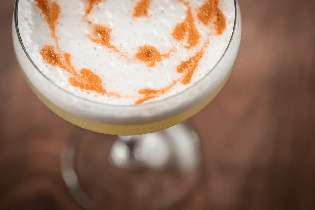 Cocktail with white creamy foam and swirled reddish-orange powder garnish.
