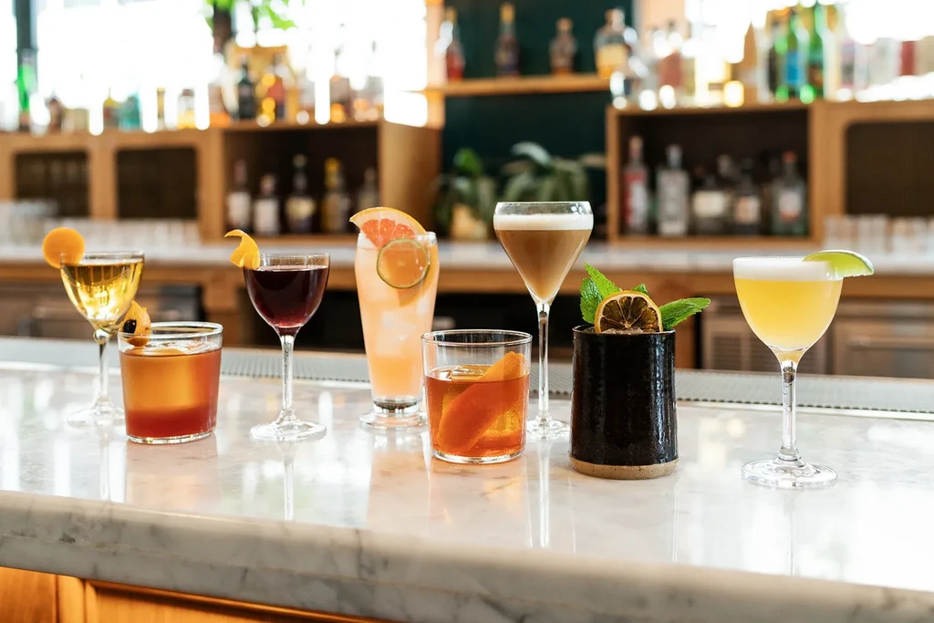 Seven different garnished cocktails on a white marble bar.