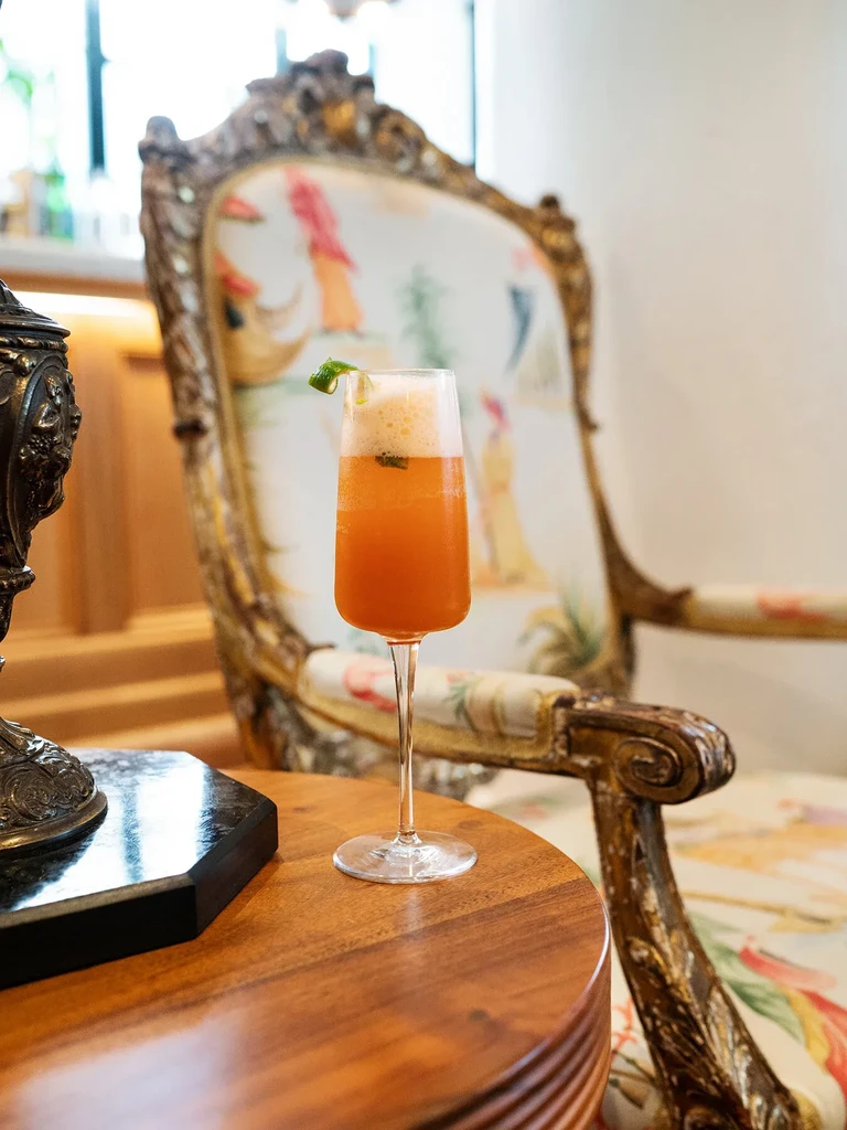 Orange drink with lime peel on wooden table, near ornate armchair.
