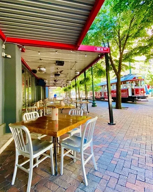 Sunny restaurant patio with red awning and vintage streetcar outside.