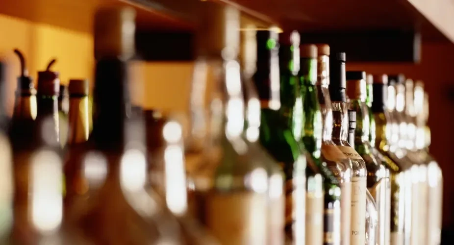 Warm-lit bar shelf displaying clear, green, and amber bottles.