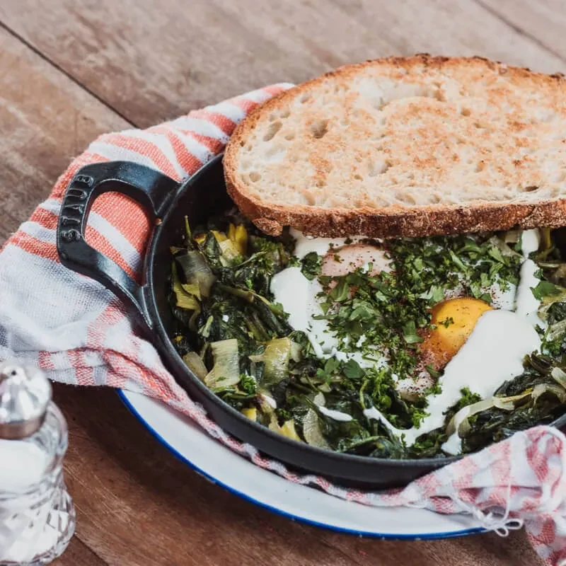 Close-up: Cast iron skillet with eggs, greens, bread, and herbs.