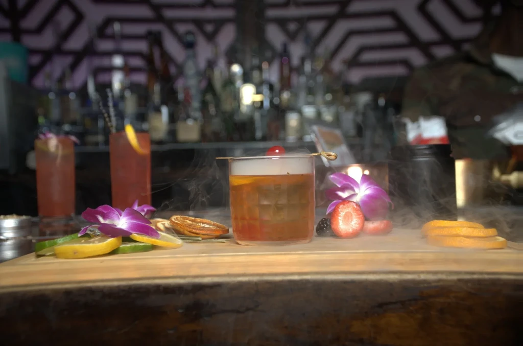 Central smoky cocktail with fruit and floral garnishes on bar.