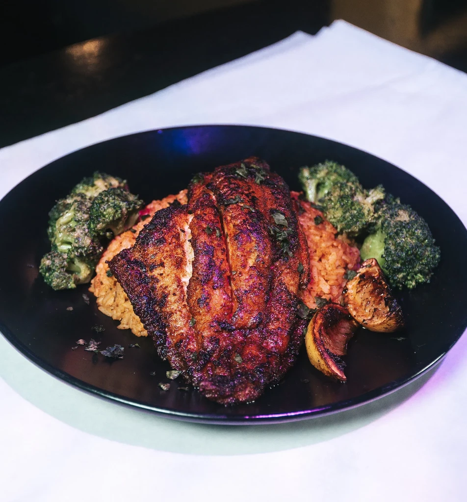 Blackened fish, reddish rice, broccoli, and charred lemon on dark plate.