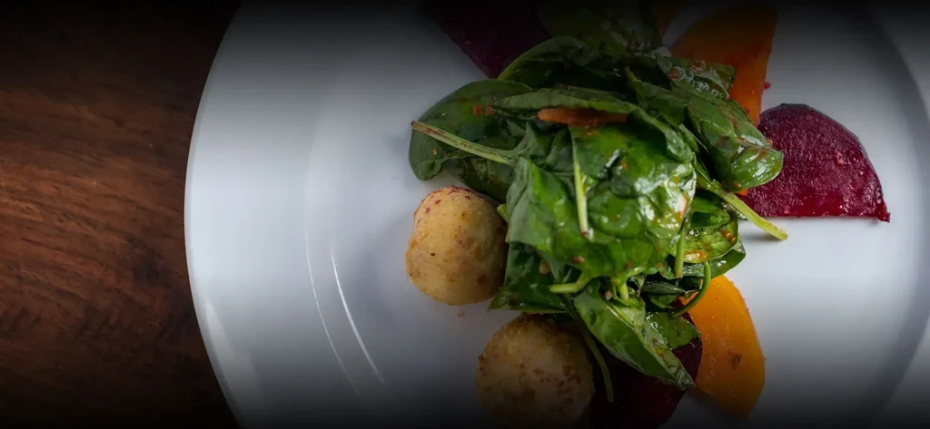 Plated spinach salad, croquettes, and root vegetables on dark wood.