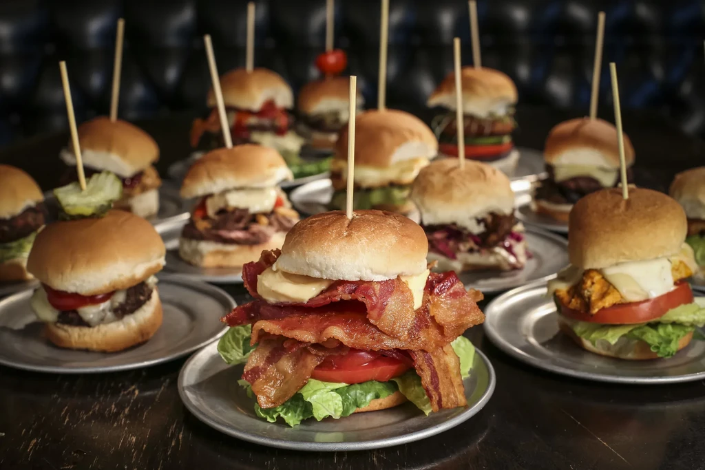 Close-up of numerous mini-burgers with diverse toppings on silver plates.