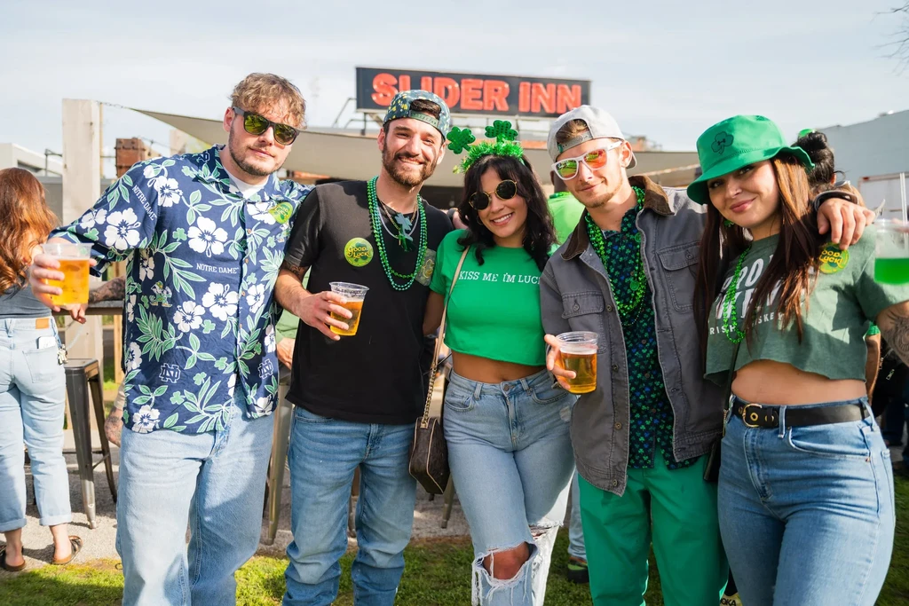 Five smiling young adults in green celebrating St. Patrick's Day outdoors.