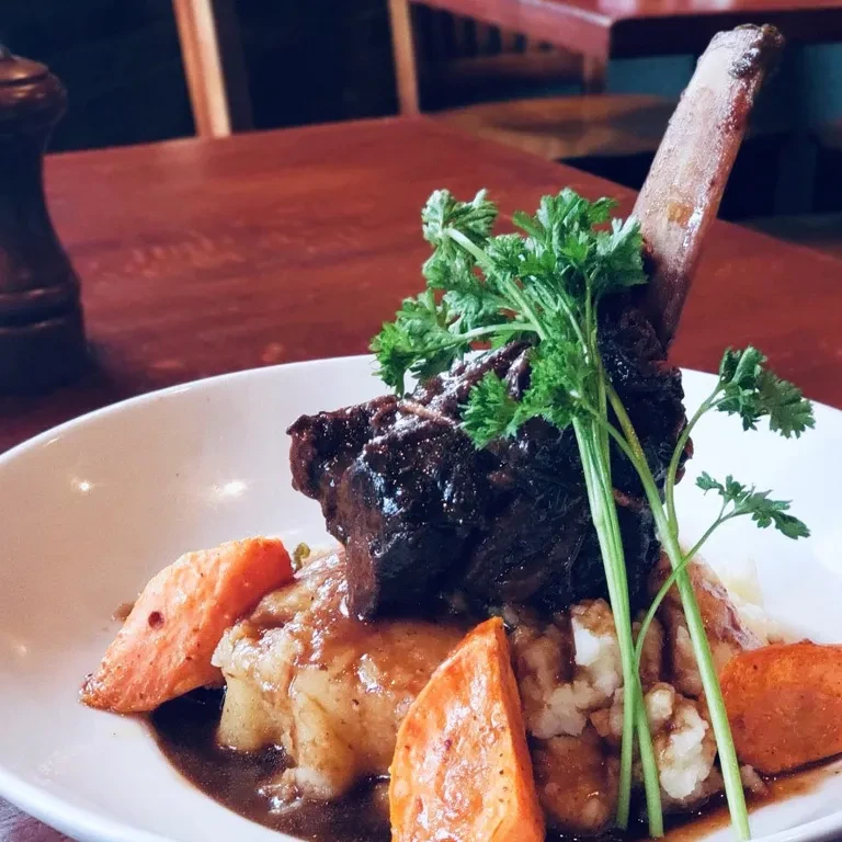Plated braised short rib with bone, mashed potatoes, carrots, and parsley.