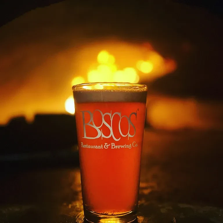 Boscos pint glass with amber beer, foam, warm blurred background.