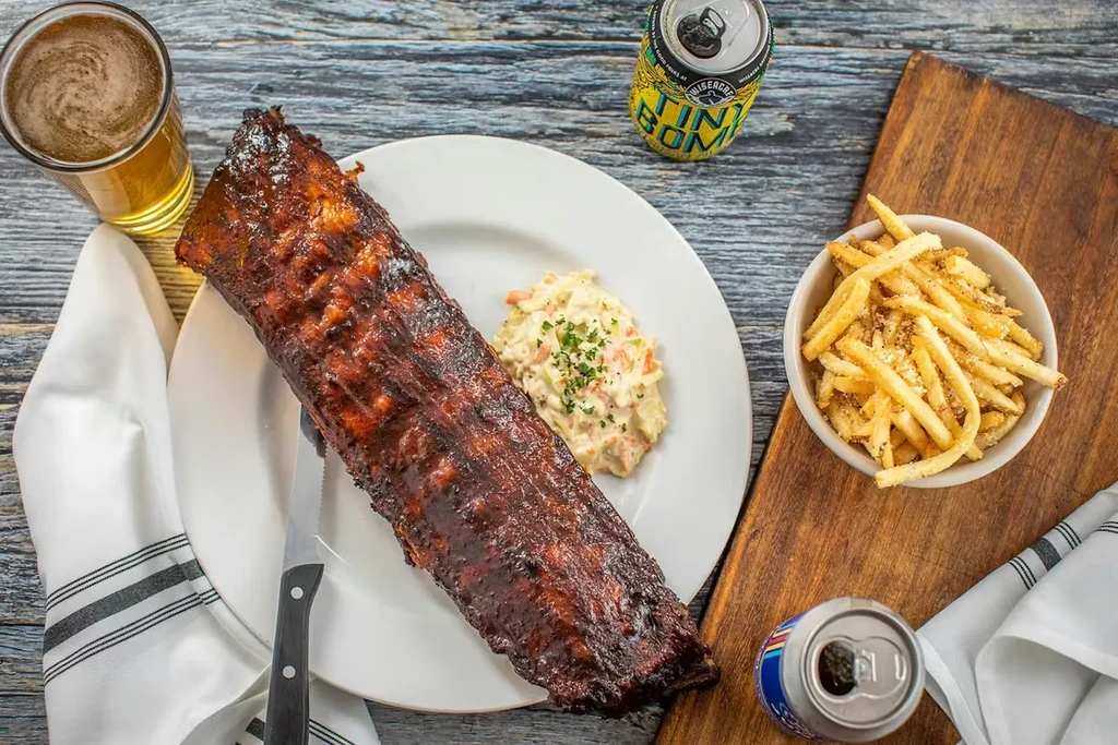 Barbecued ribs, coleslaw, fries, and drinks on a wooden table.