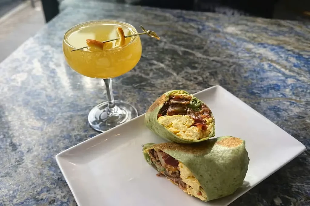Halved breakfast burrito and yellow cocktail on a stone counter.